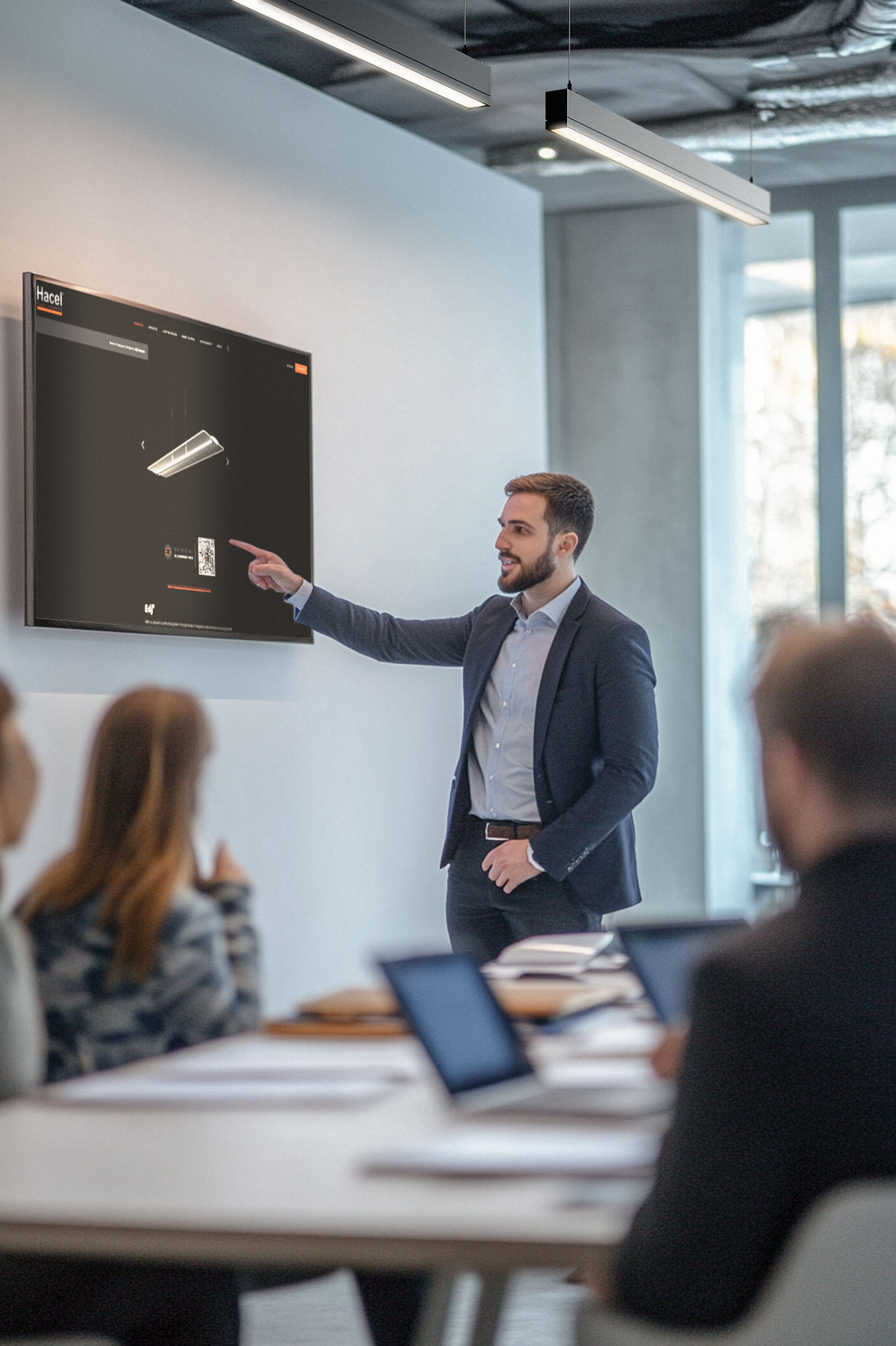 A photo of an office worker presenting on a digital screen to his colleagues in the conference room, showing Hacels product web page. He's pointing at the screen displaying a Hacel QR code that leads to an immersive AR experience.